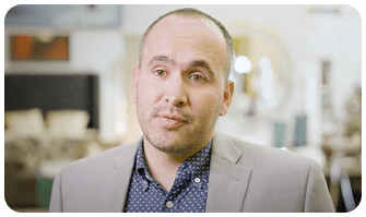 A man in a gray suit and blue polka dot shirt stands indoors, with blurred furniture and decor in the background.