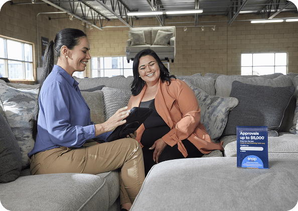 Two women sitting on a showroom couch reviewing a tablet, with Snap Finance signage offering up to $5,000 approval.