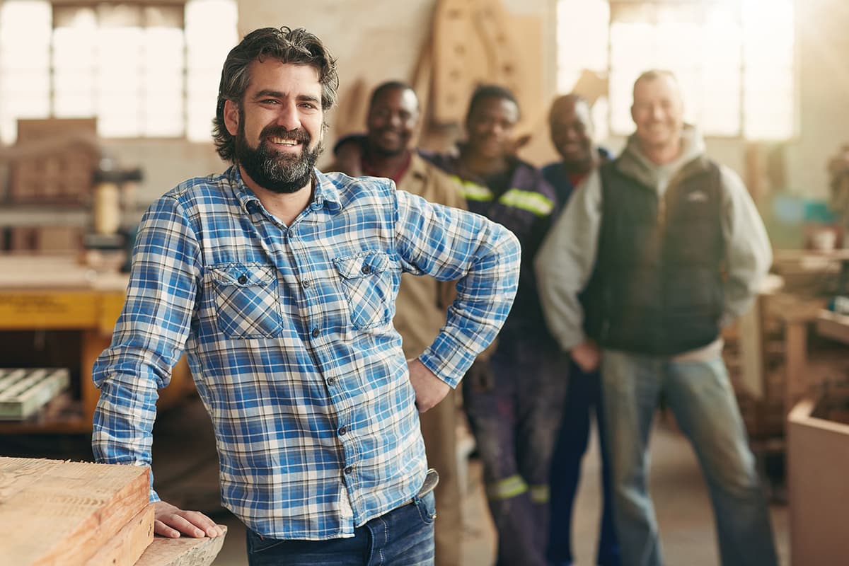 Carpenter or woodworker in blue plaid shirt smiling confidently in workshop, with team members standing in background