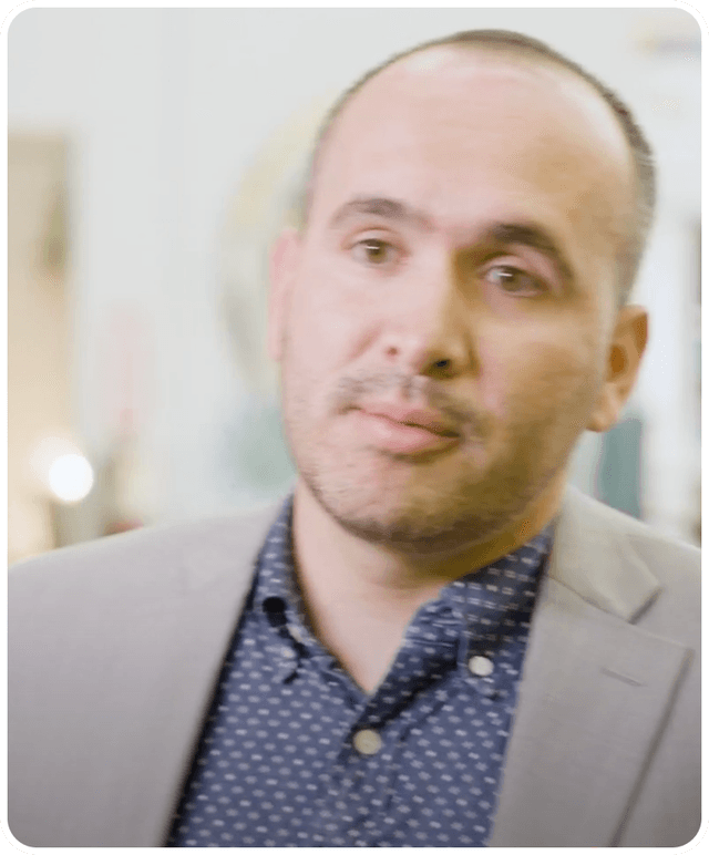 A man with short hair and a beard, wearing a polka dot shirt and gray blazer, appears in a softly blurred indoor setting.